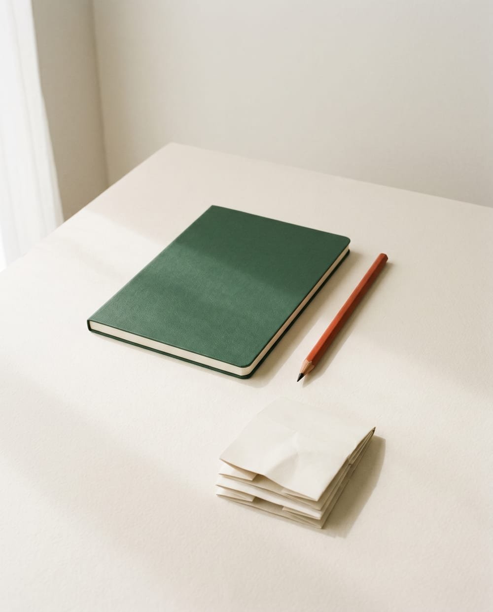 A green notebook, a terracotta pencil and folded paper swatches on a cream studio desk
